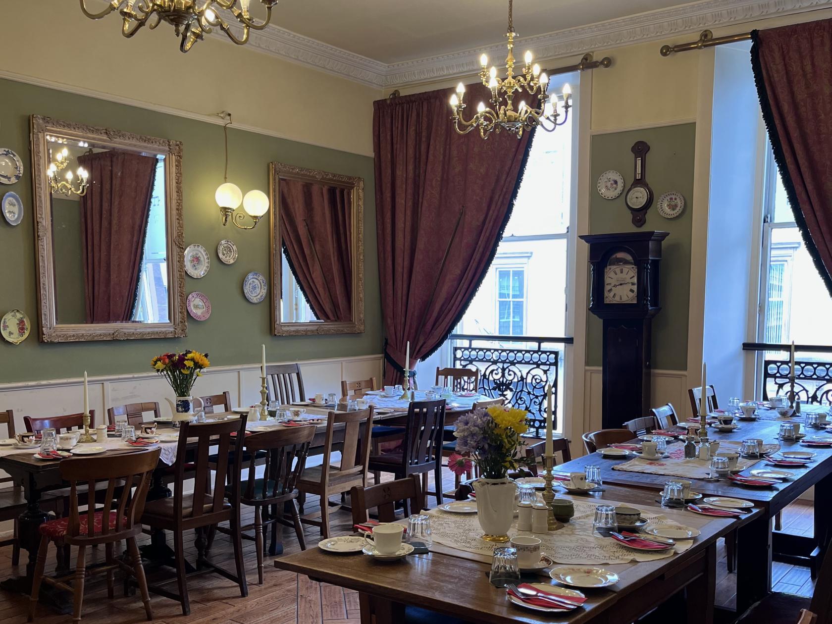 A dining room with vintage wooden tables, floral china, tall windows with drapes, and decorative wall mirrors at The Butterfly and The Pig.