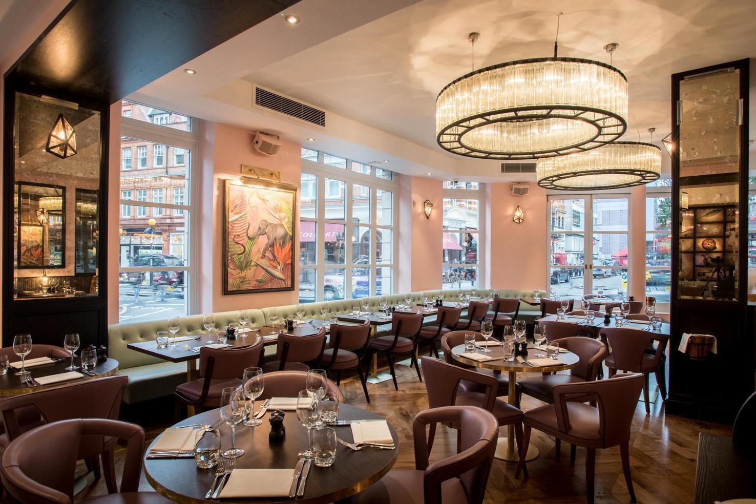 A cocktail bar with round tables, blush seating, large windows, and crystal chandeliers at The Botanist Sloane Square.