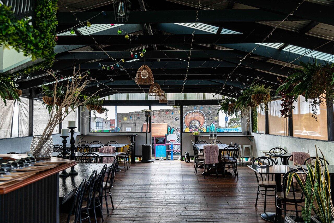 A covered outdoor dining area with wooden floors, hanging plants, and round tables at The Den Cafe and Bar.