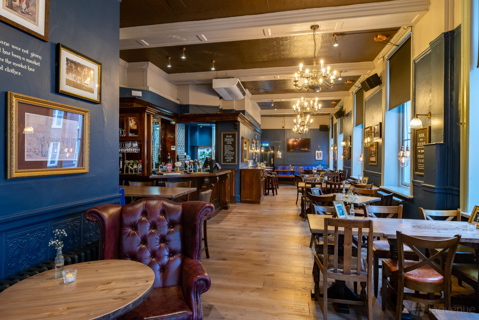 A pub dining room with wooden floors, chandeliers, leather armchairs, and framed artwork at The Woodins Shades.