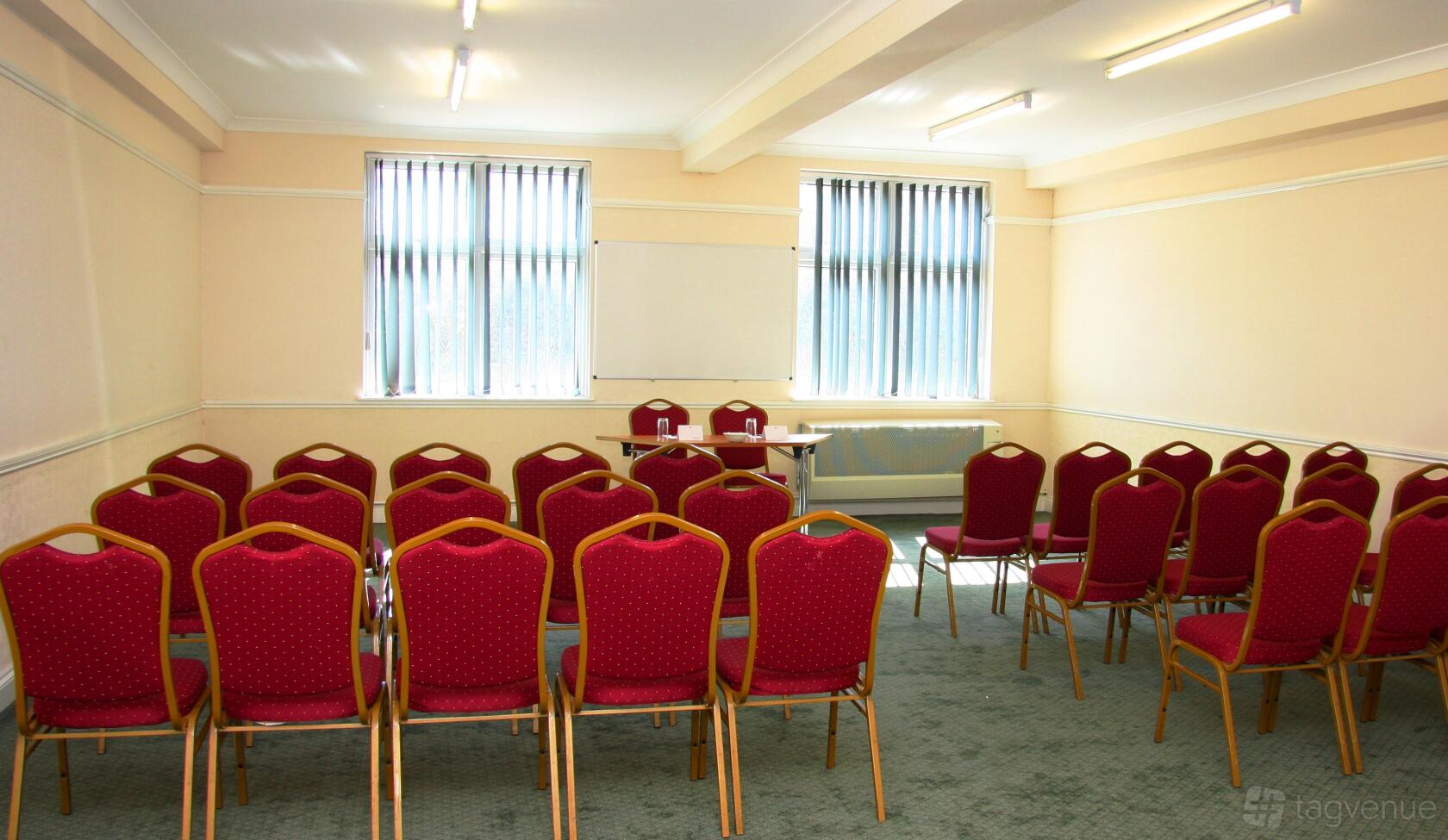 A meeting room with red cushioned chairs in rows, a table at the front, and large windows at The Thurrock Hotel.