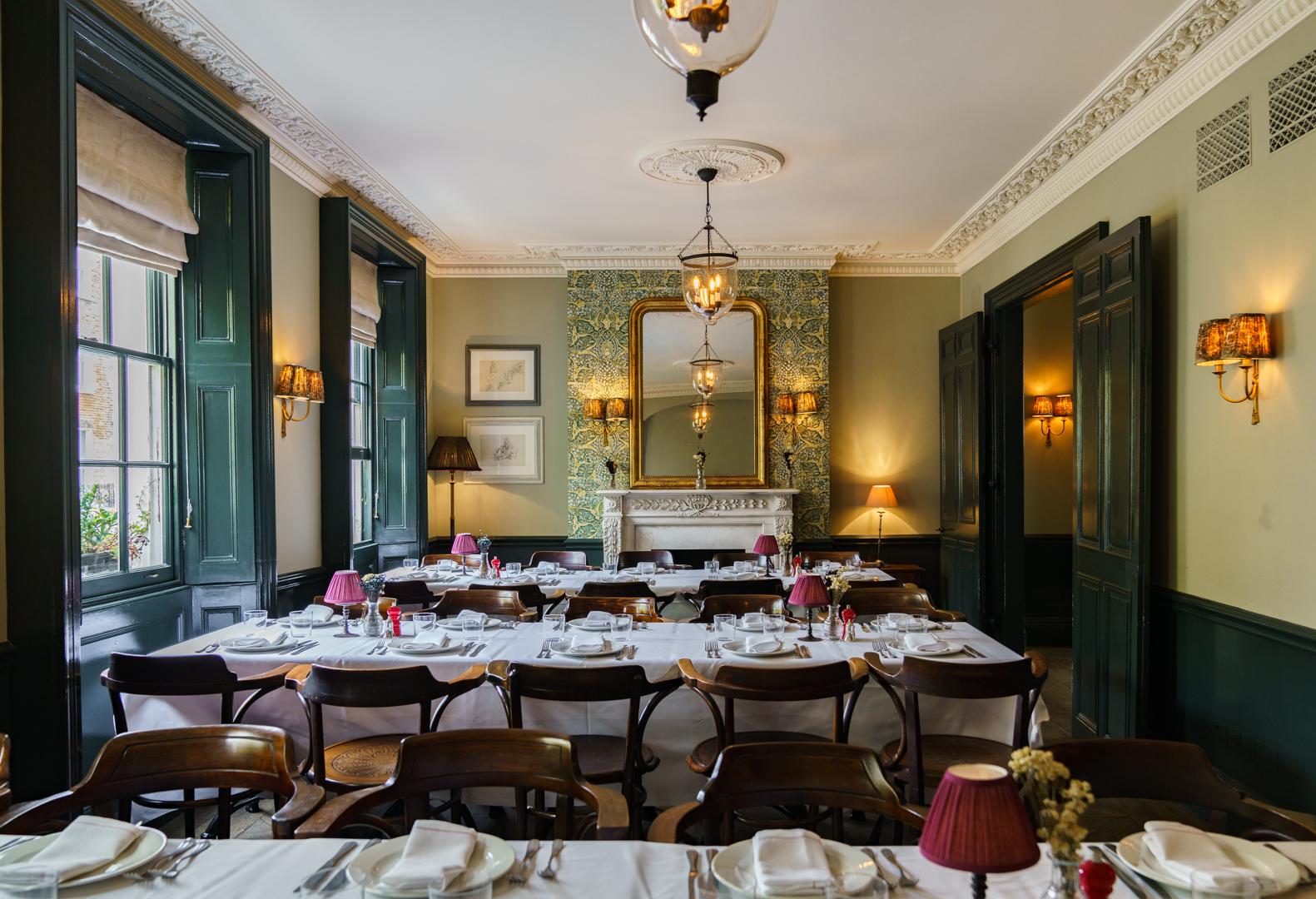 A pub dining room with dark wood chairs, white tablecloths, and a fireplace with a decorative mirror at The Thomas Cubitt.