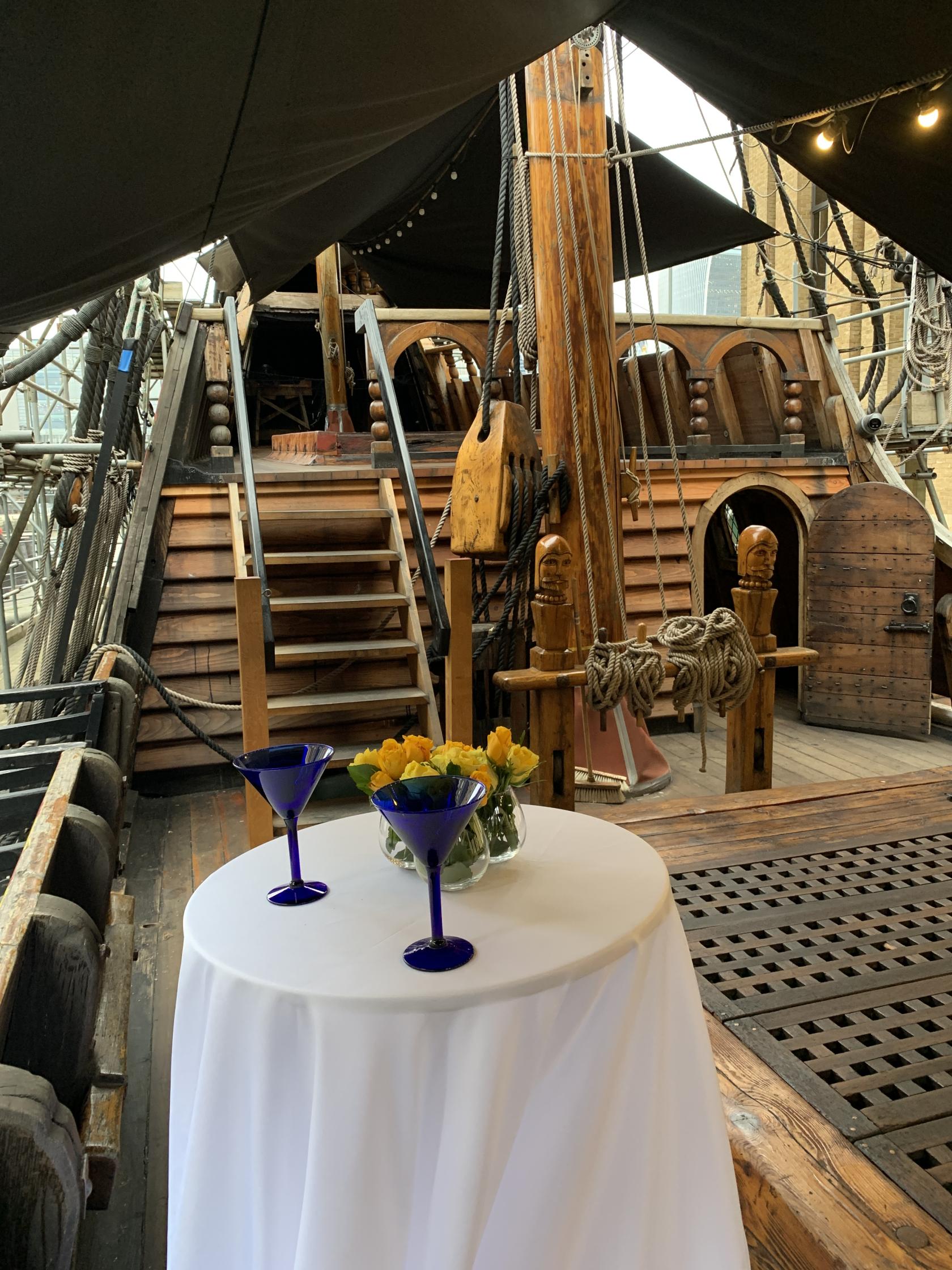 A ship museum with exposed wooden decks, nautical ropes, and a table set with blue glasses and yellow flowers at The Golden Hinde.
