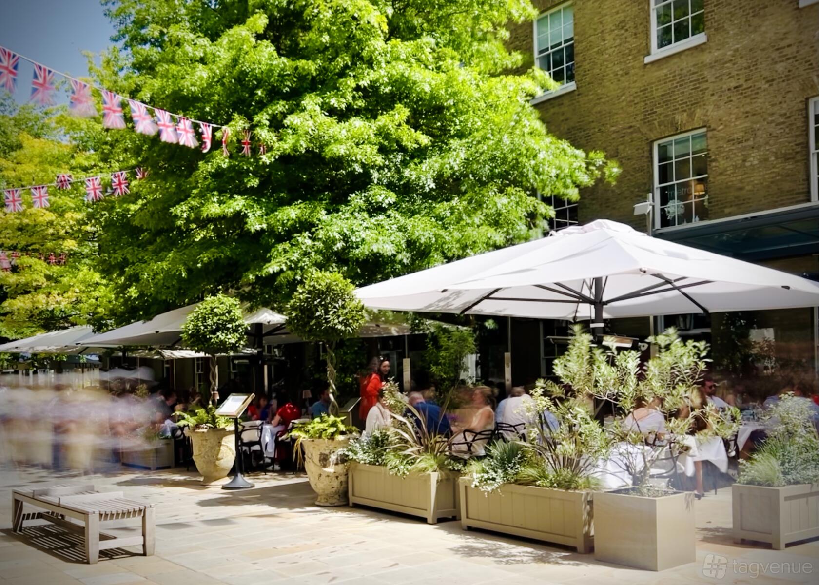 An outdoor restaurant terrace with large umbrellas, potted plants, and diners under trees at Manicomio Chelsea Restaurant.