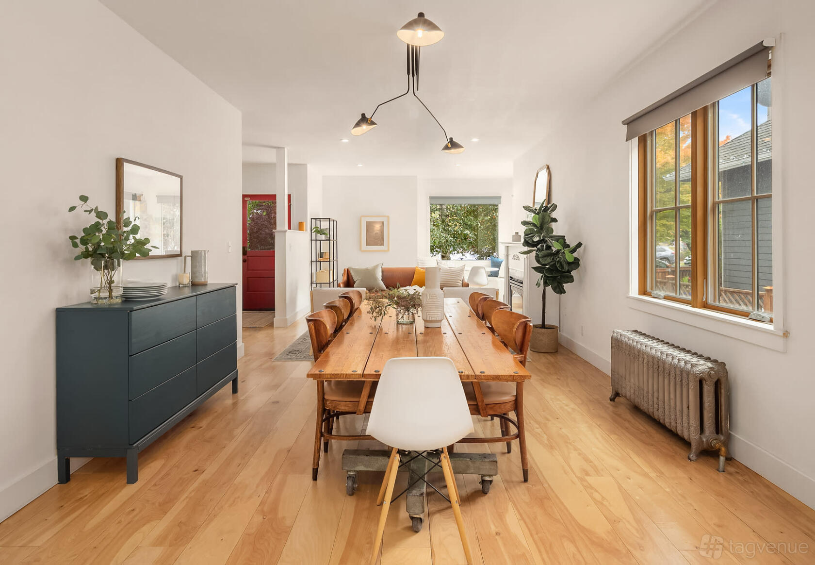 A house interior with a long wooden dining table, light wood floors, large windows, and leafy plants at Light Filled Home.