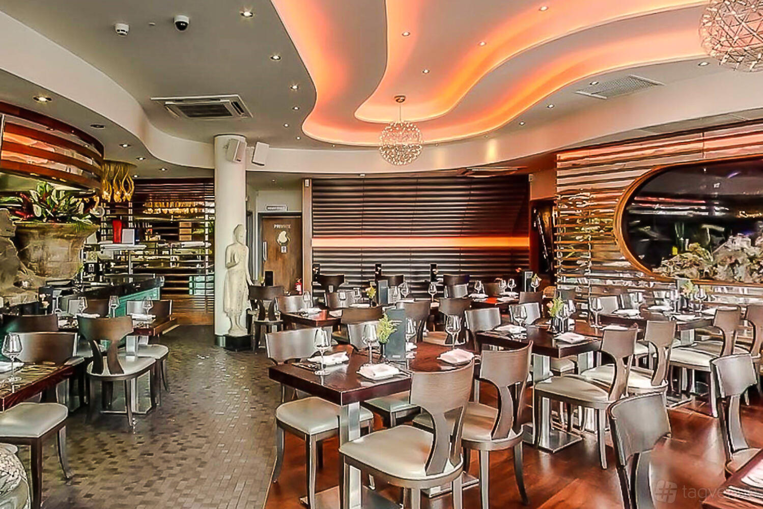 A restaurant with warm ceiling lighting, curved wood paneling, and neatly arranged tables at Chaophraya Birmingham.
