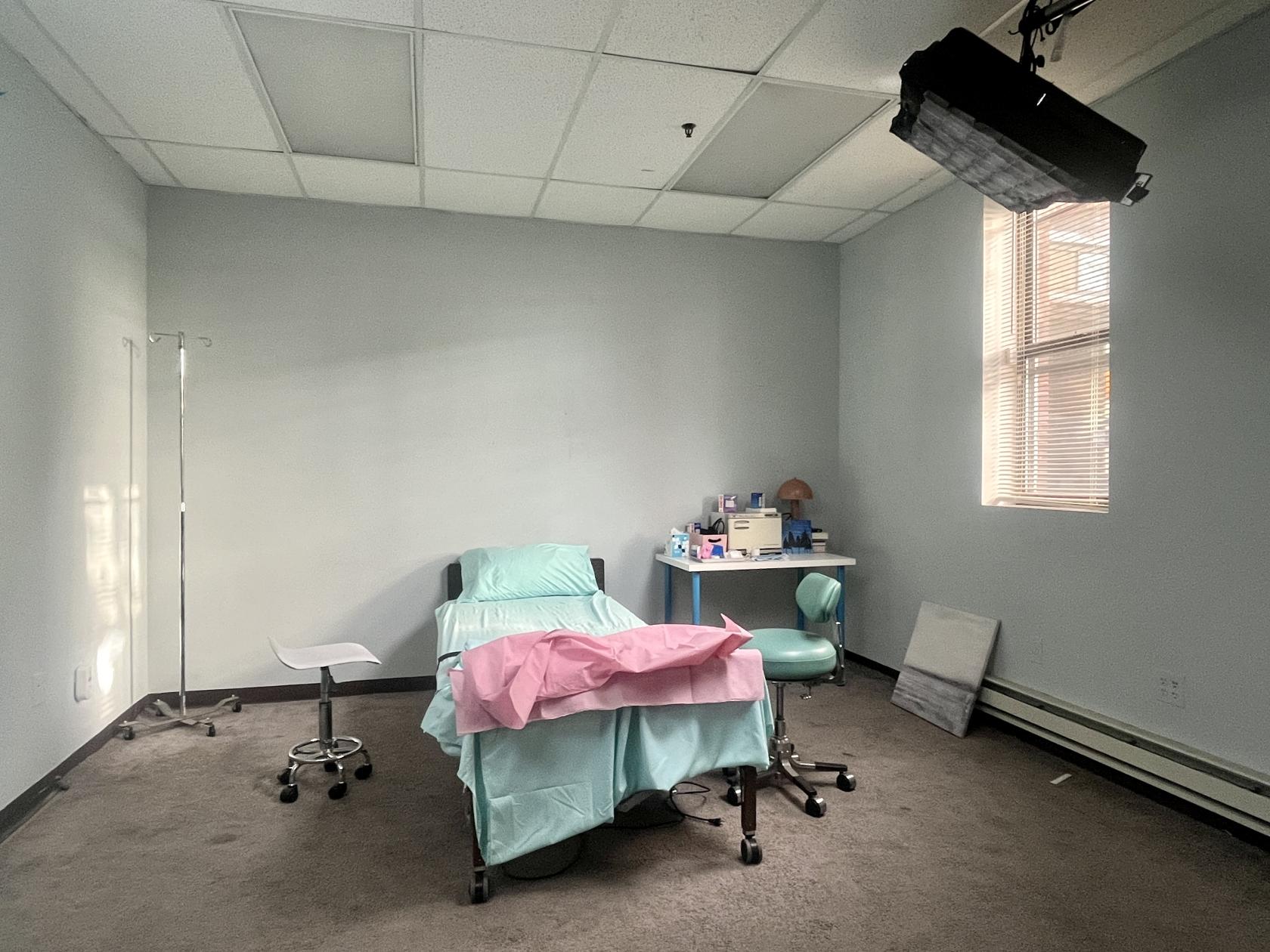 A photo studio with a hospital bed, IV stand, and medical props in a room with natural light at Hospital Room: Doctor's Office.