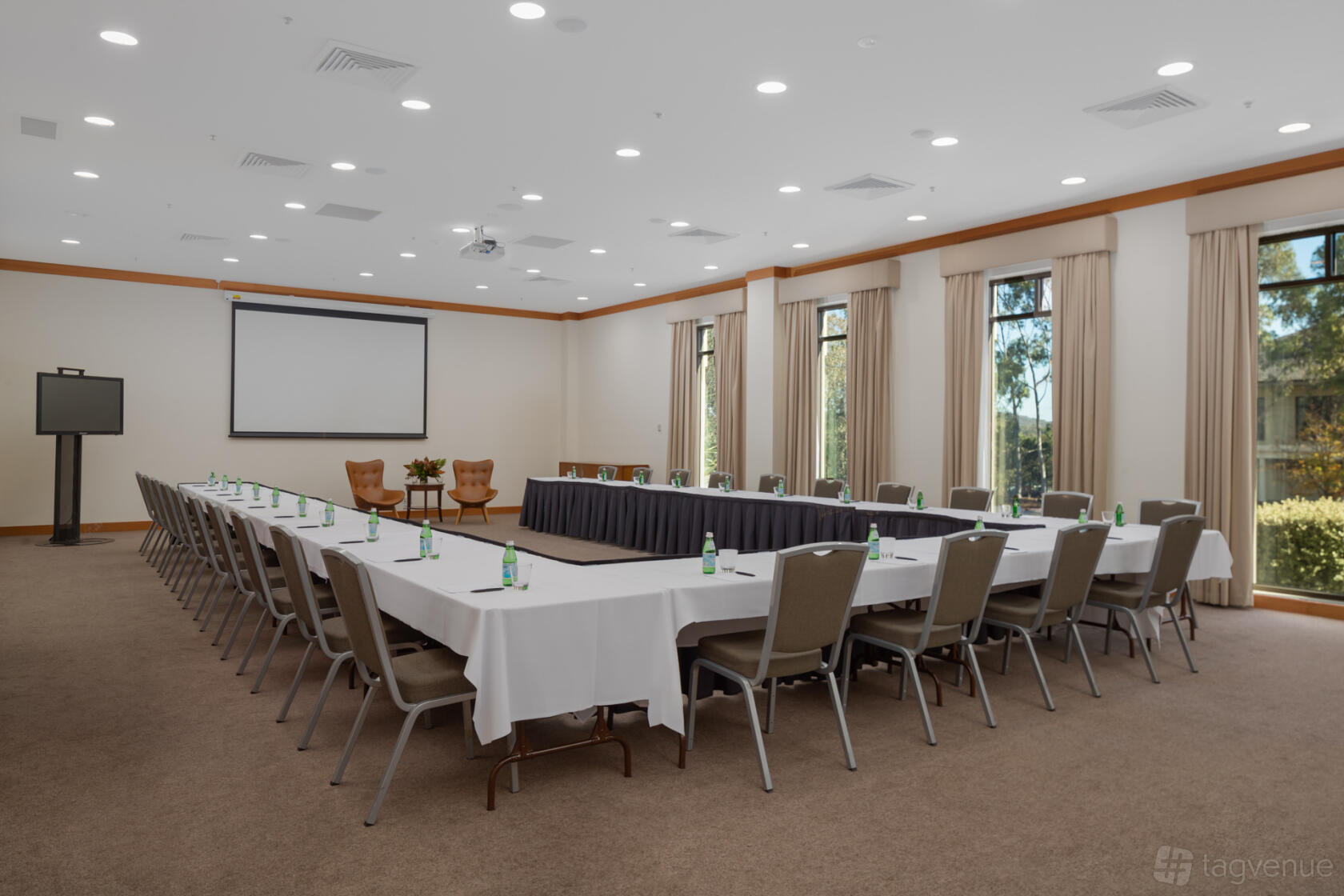 An event space with U-shaped tables, bottled water, large windows, and a projection screen at Yarra Valley Lodge.