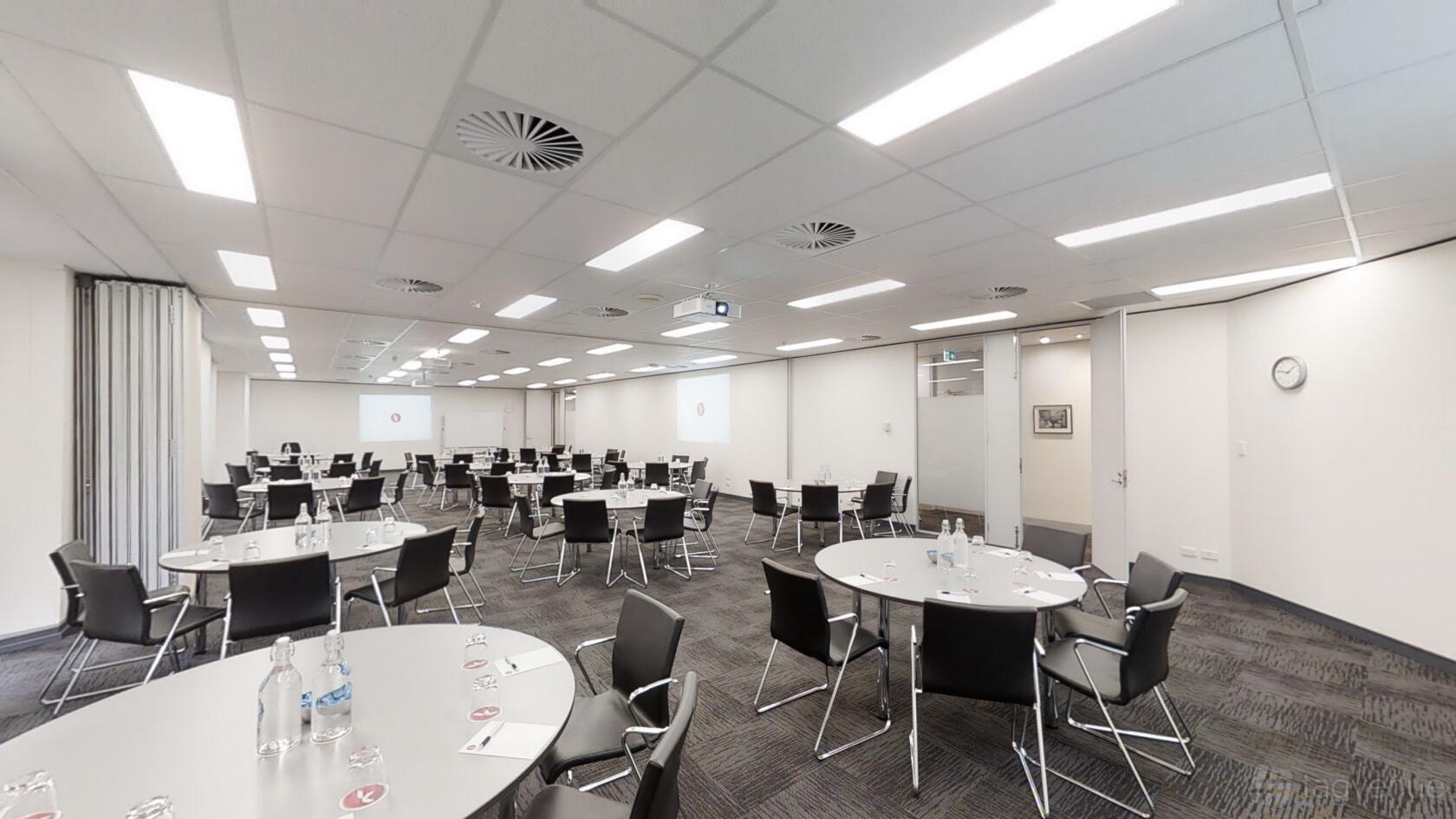 A conference centre room with round tables, black chairs, dual projection screens, and ceiling lights at Karstens Perth.