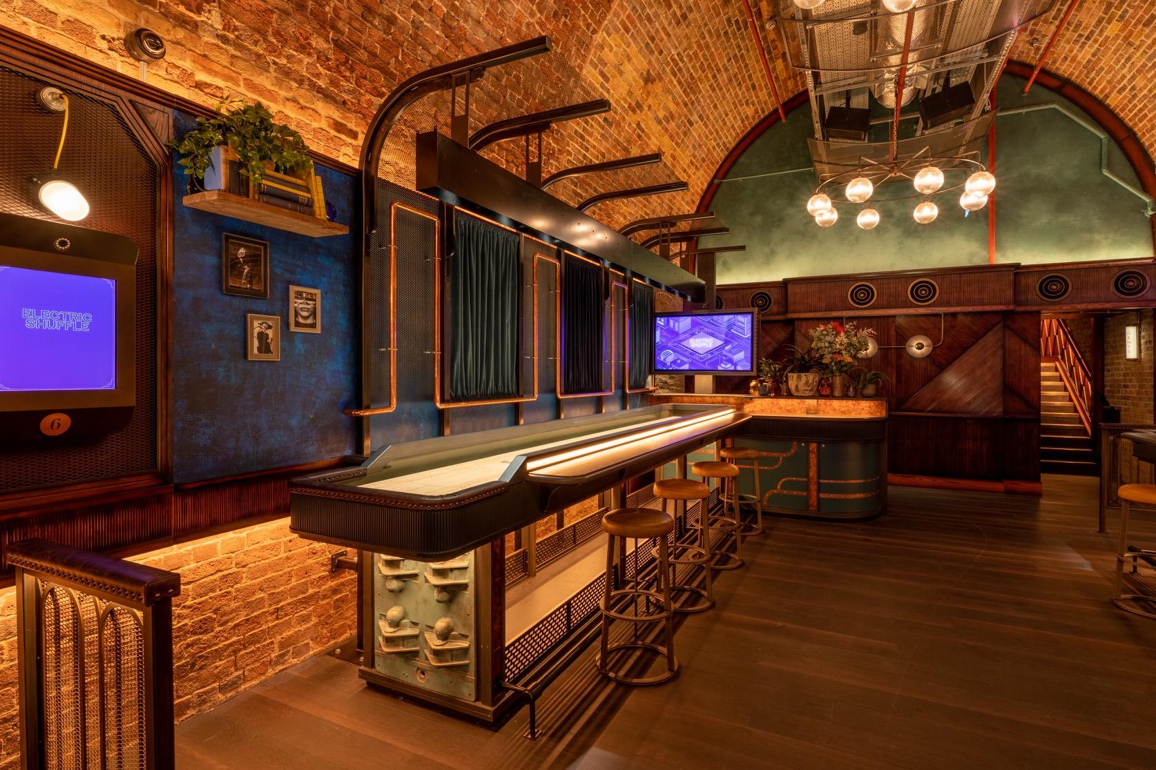 An event space in a cocktail bar with exposed brick arches, shuffleboard table, and bar seating at Electric Shuffle London Bridge.