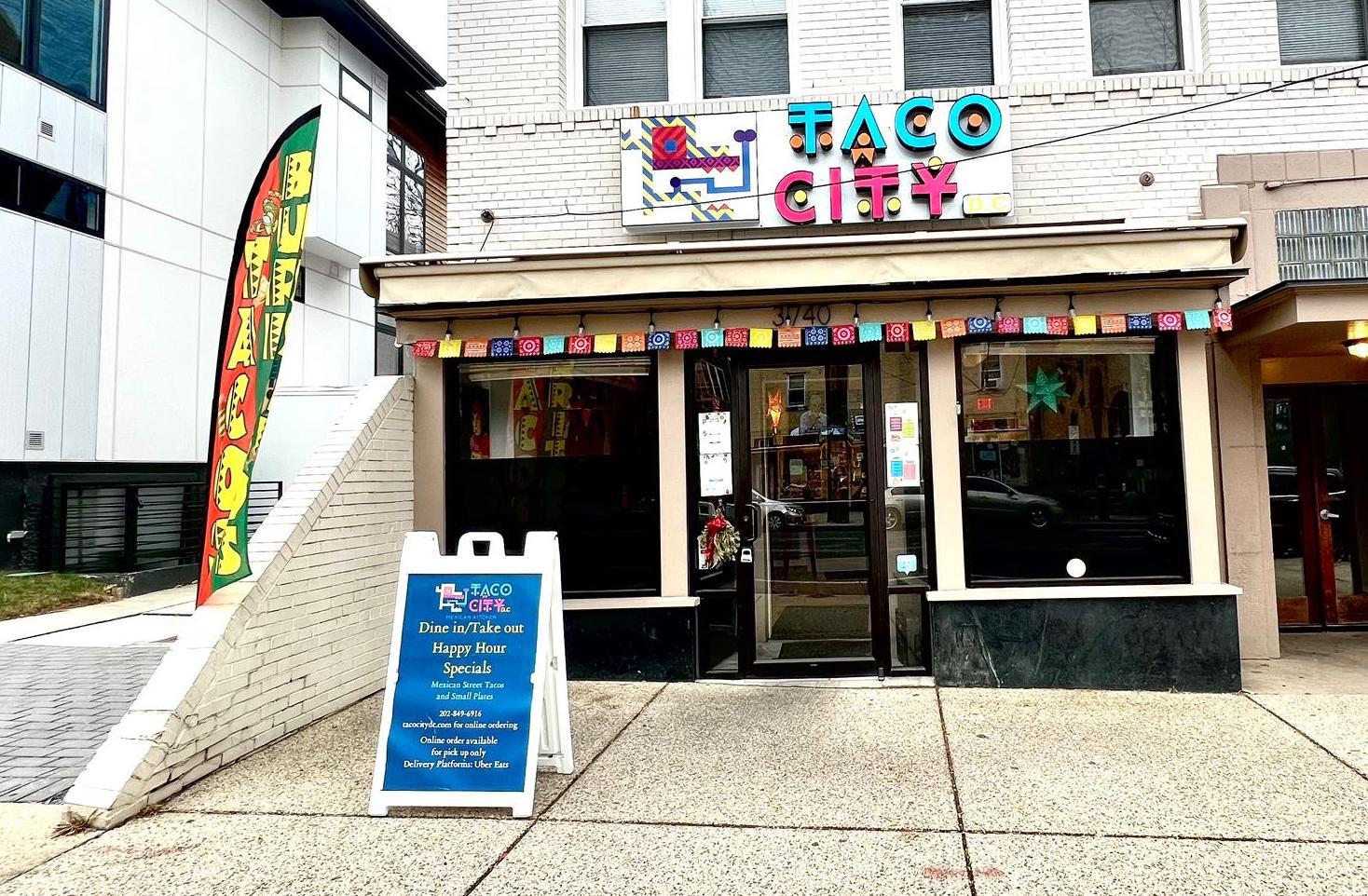 A restaurant entrance with a colorful sign, glass front windows, and sidewalk signage at Taco City DC.