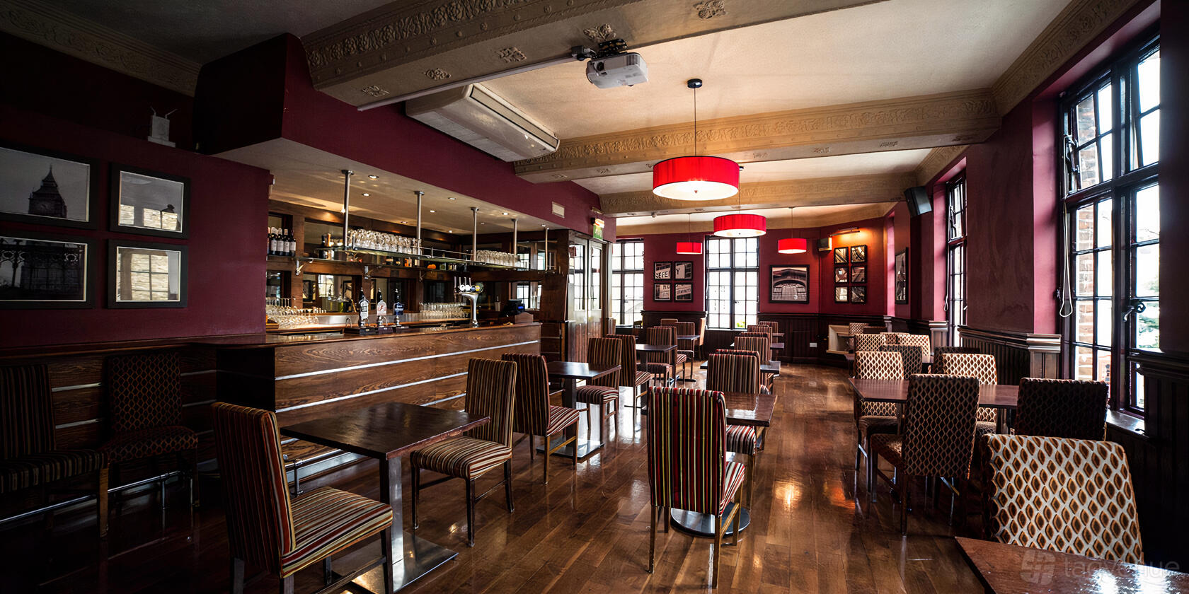 A bar in a pub with striped upholstered chairs, wood floors, and red pendant lighting at The Barley Mow.