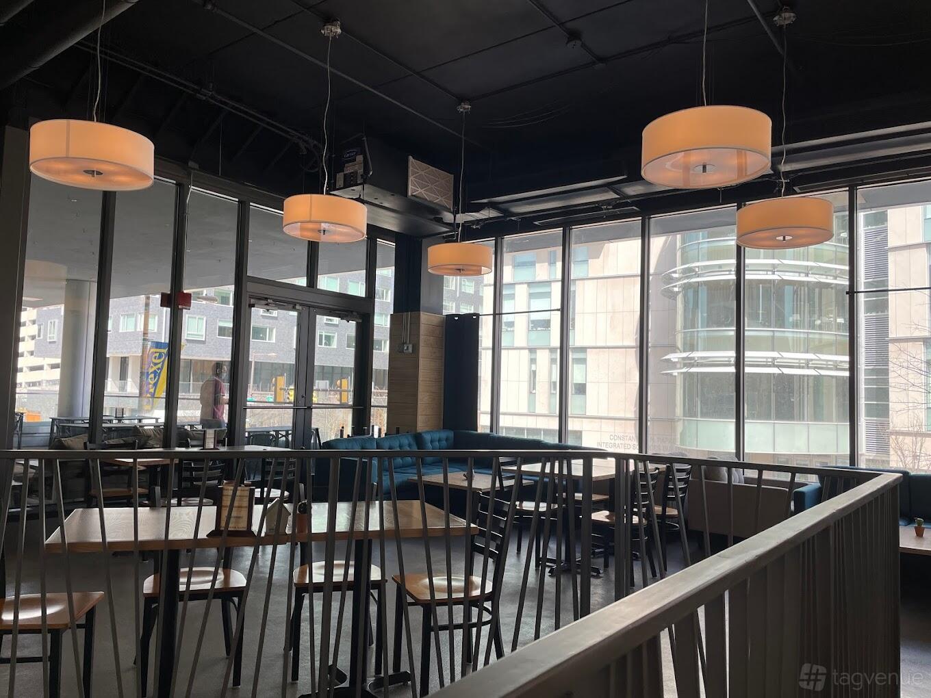 A restaurant with floor-to-ceiling windows, dark ceiling, round pendant lights, and wooden tables at The Board and Brew.