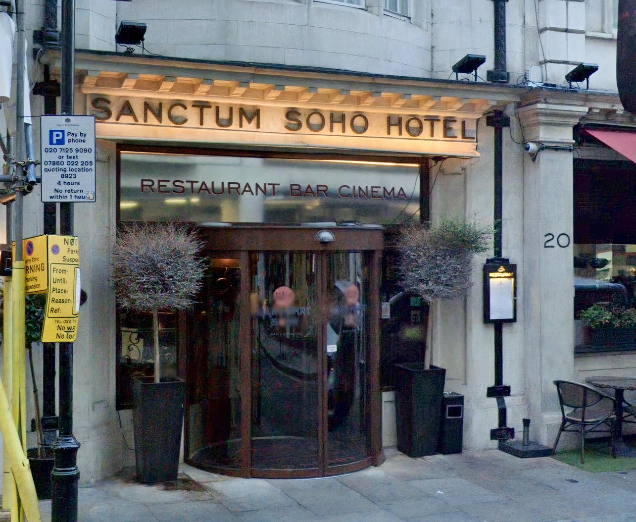 A hotel entrance with a revolving glass door, illuminated signage, and potted plants at Karma Sanctum Soho Hotel.