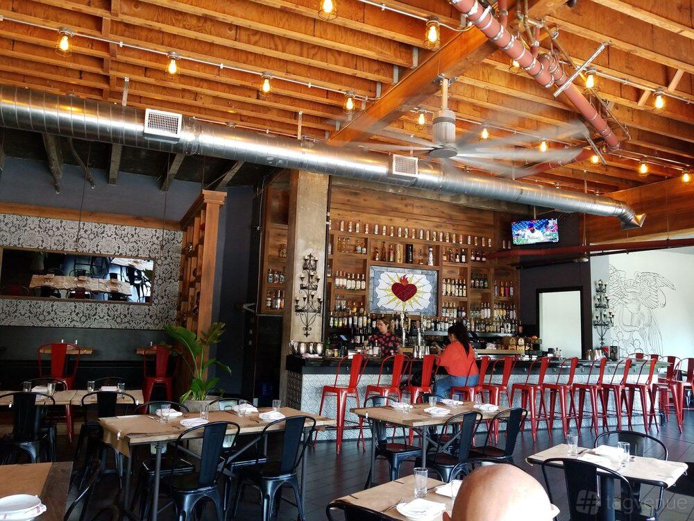 A restaurant with exposed wood ceilings, hanging string lights, and a colorful bar with red stools at Mi Corazon.