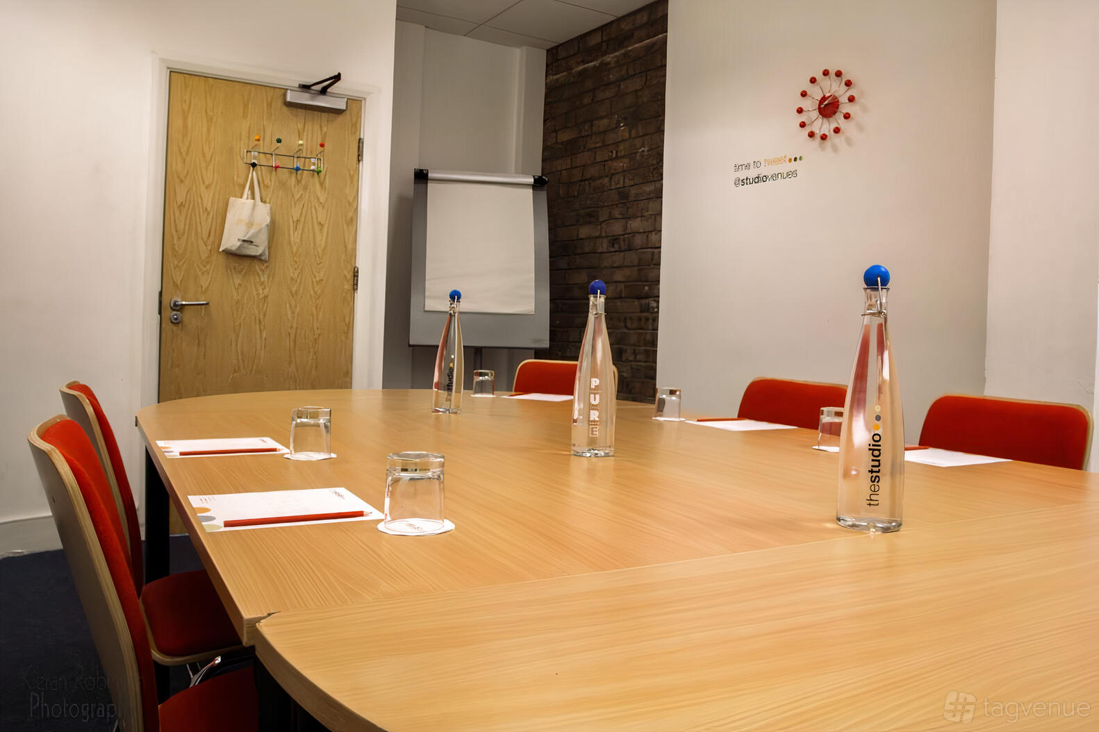 A meeting room with a wooden oval table, red chairs, exposed brick wall, and bottled water at The Studio Birmingham.