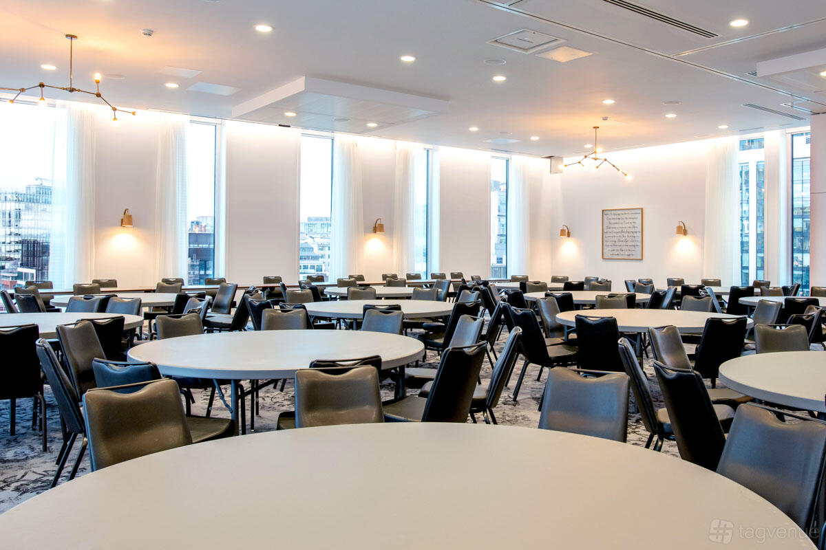 A hotel hall with round tables, black chairs, large windows, and modern ceiling lights at voco Manchester.