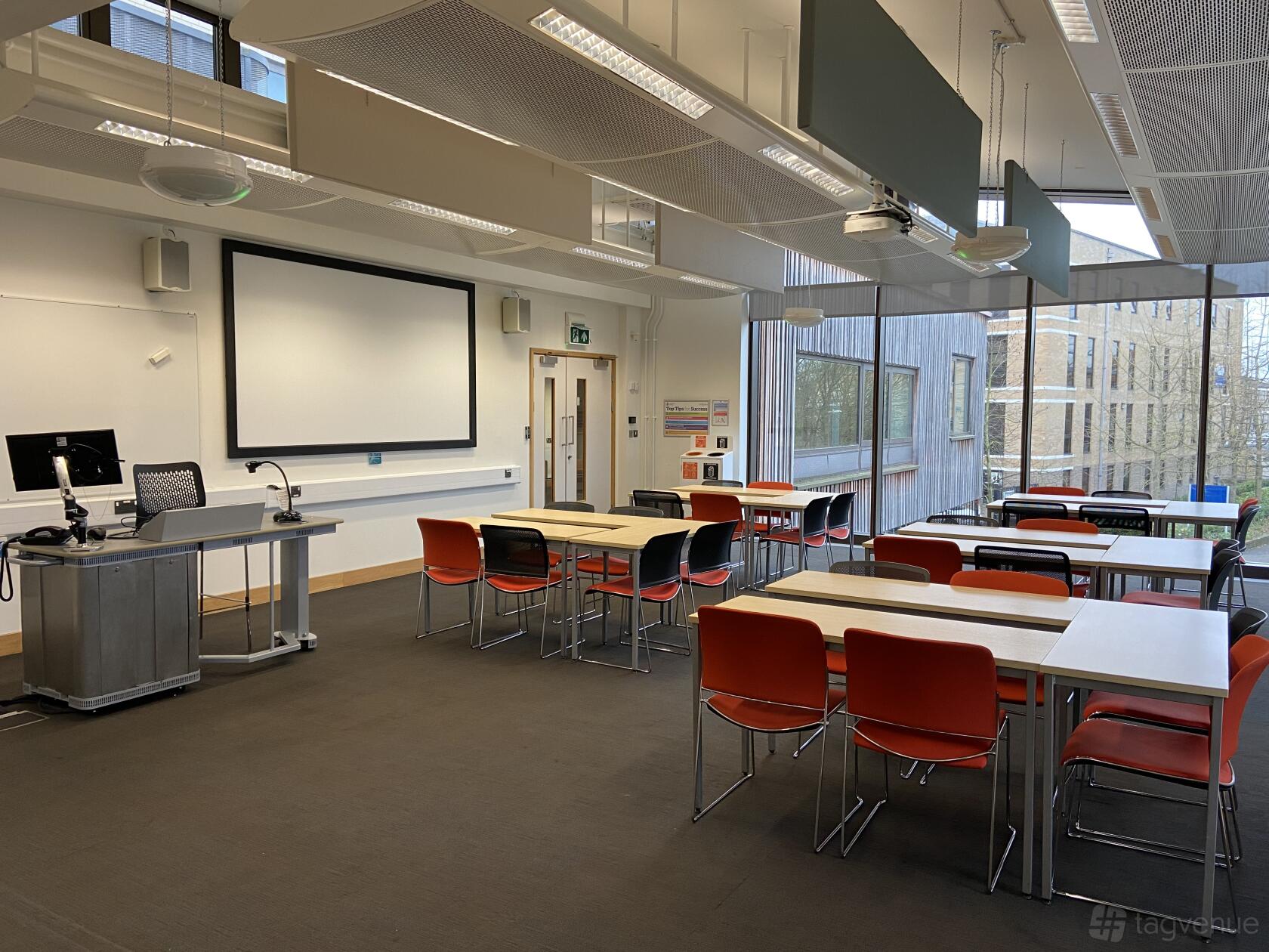 A classroom with rows of desks and red chairs, projector screen, and large windows at ARU Venue Hire - Cambridge.