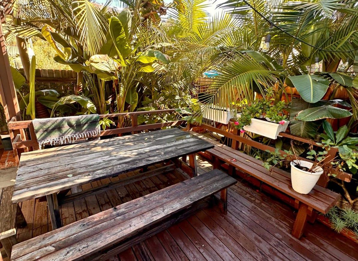 A restaurant patio with wooden picnic tables surrounded by lush tropical plants at Cottage Home with Hawaii Vibes.