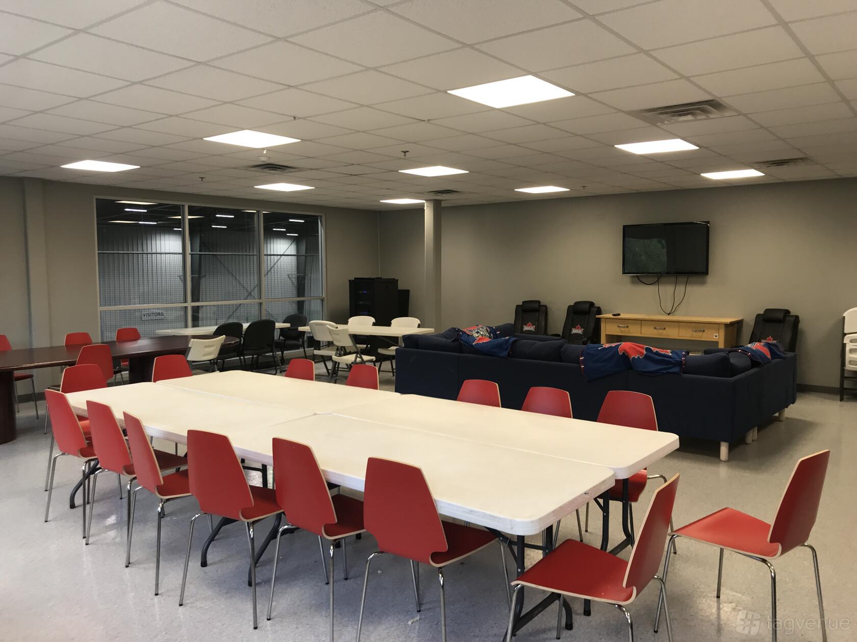 A meeting room with rectangular tables, red chairs, a wall-mounted TV, and floor-to-ceiling windows at Toronto Rock Athletic Centre.