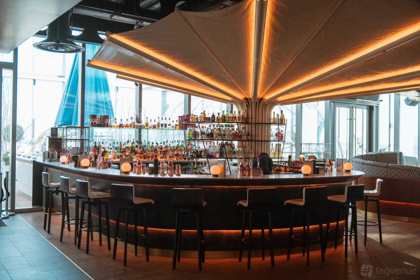 A restaurant with a circular central bar, high-backed stools, backlit shelving, and floor-to-ceiling windows at 20 Stories.