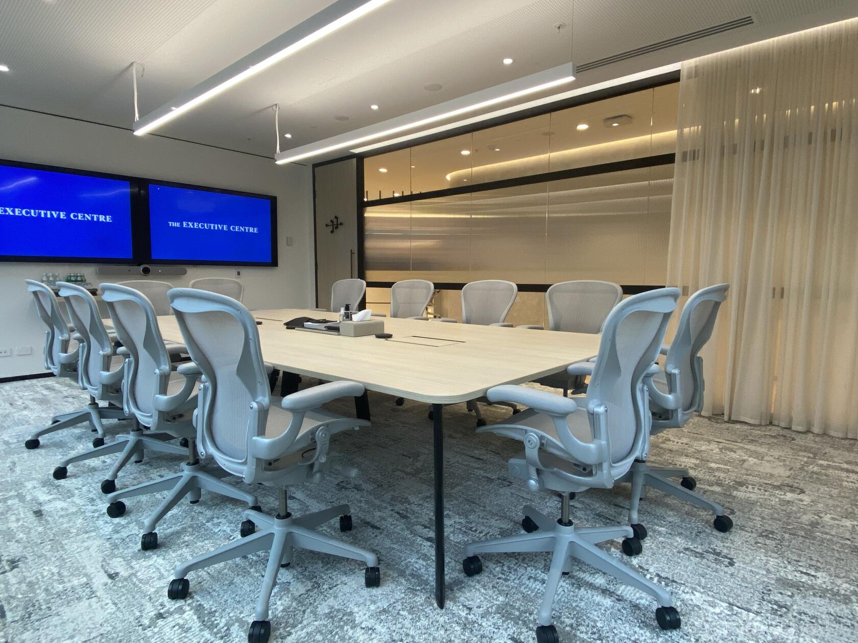 A boardroom with ergonomic chairs, dual wall-mounted screens, and a light wood table at Angel Place.