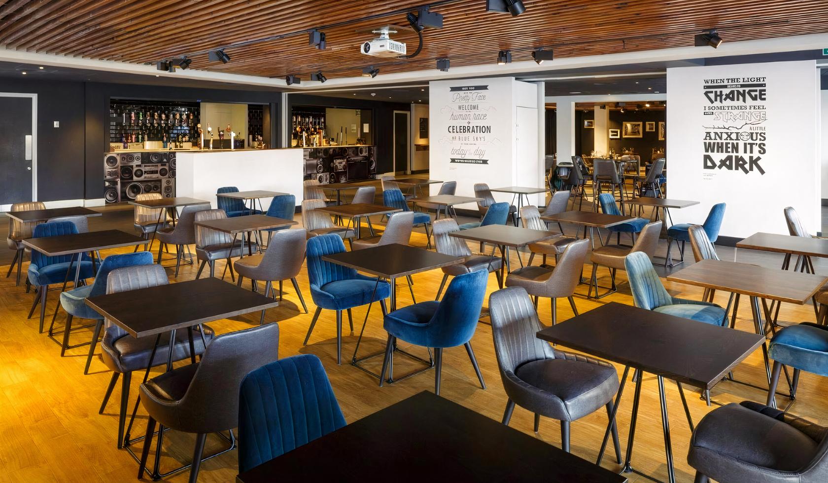 A lounge with wooden ceiling slats, a fully stocked bar, and rows of black tables with mixed upholstered chairs at first direct bank area - Leeds.