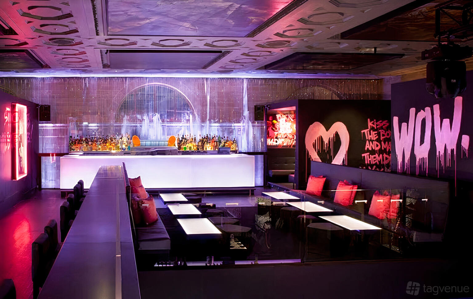 A nightclub with glowing bar, neon wall art, and cushioned booths with pink accent pillows at Ms Collins.