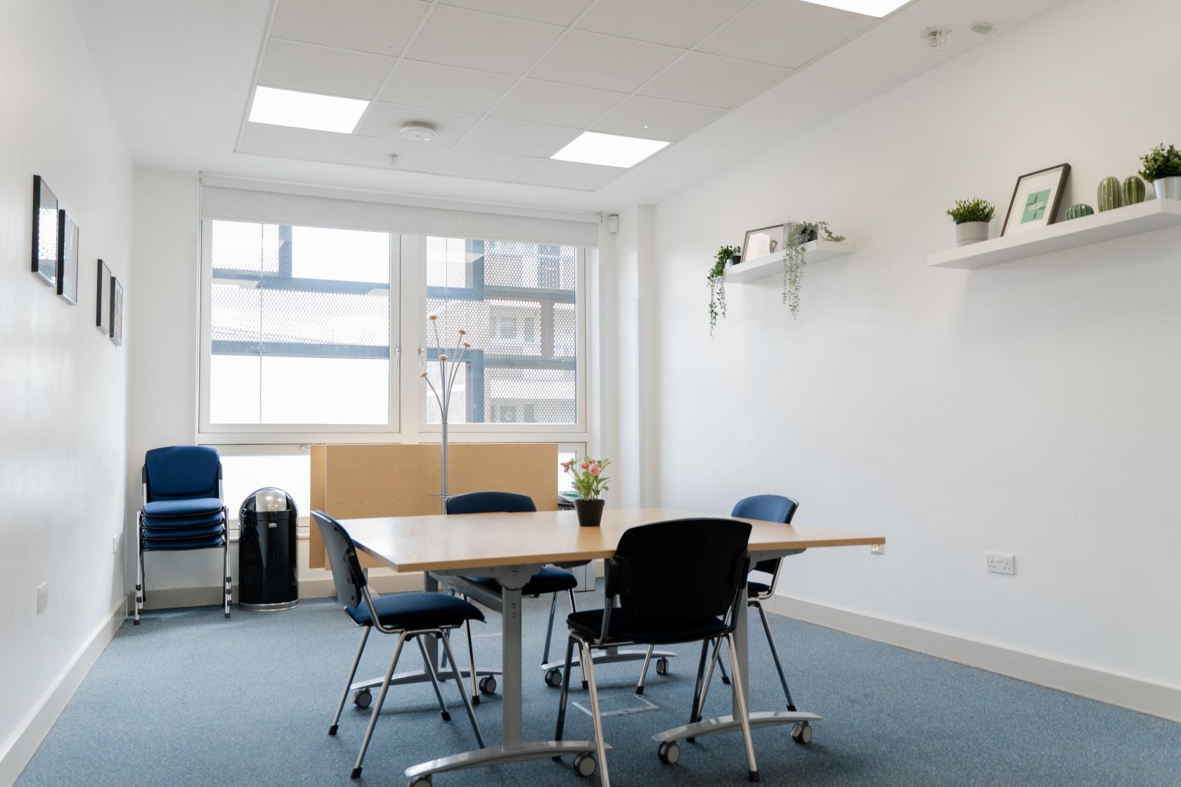 A meeting room with a rectangular table, black chairs, large windows, and wall shelves with potted plants at Deptford Lounge.