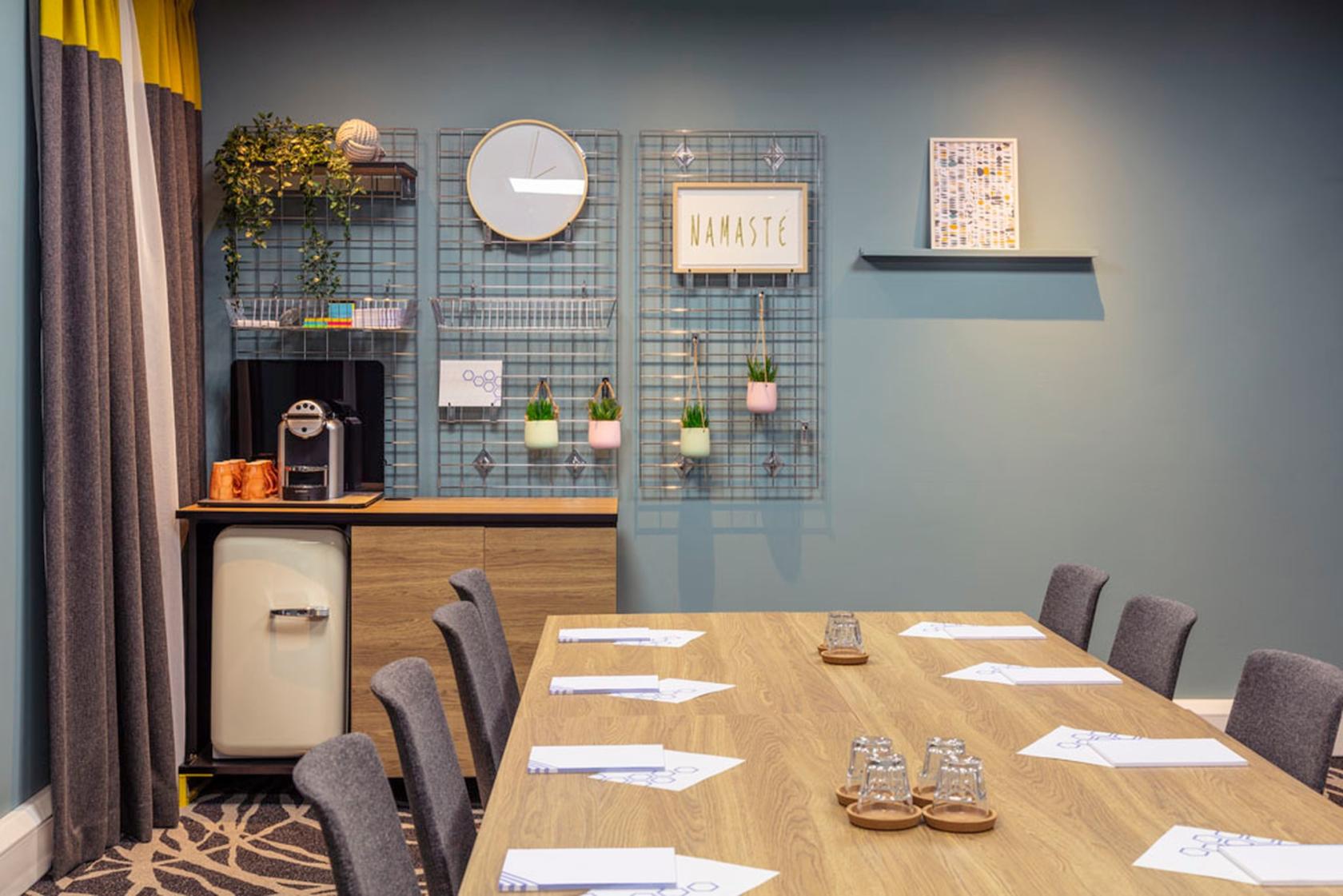 A hotel meeting room with a wooden table, gray chairs, a mini fridge, and decor including wall art and potted plants at Novotel Edinburgh Centre.