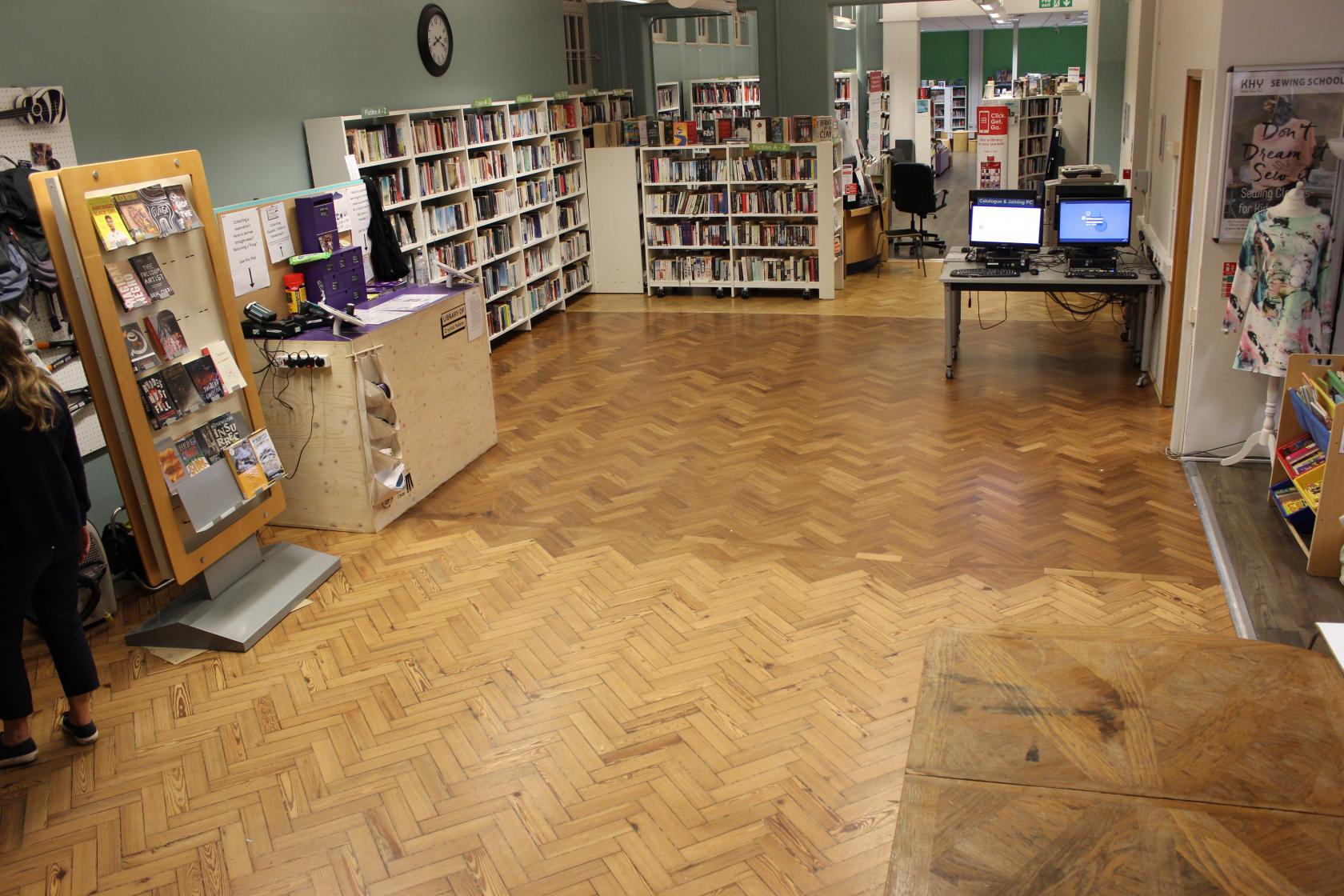 A library with parquet flooring, tall bookcases, reading areas, and self-service computer stations at The Upper Norwood Library Hub.