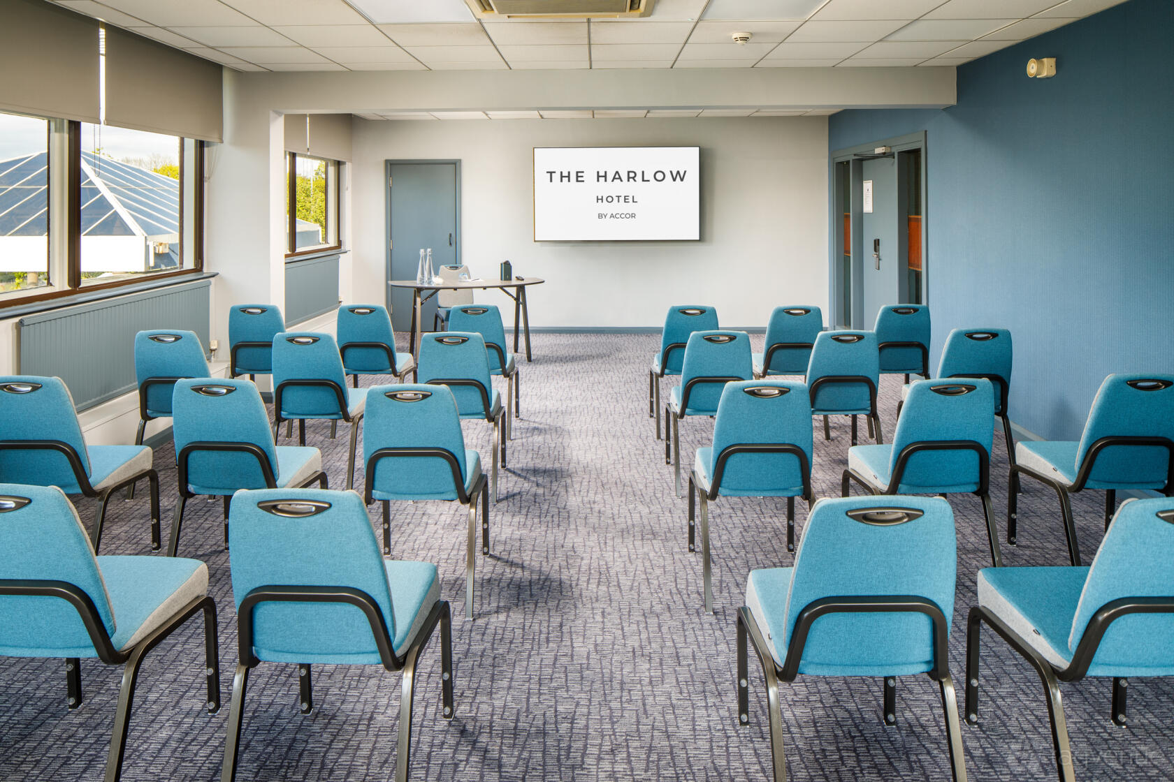 A meeting room with rows of blue chairs, a projector screen, and large windows at Harlow Hotel by Accor.