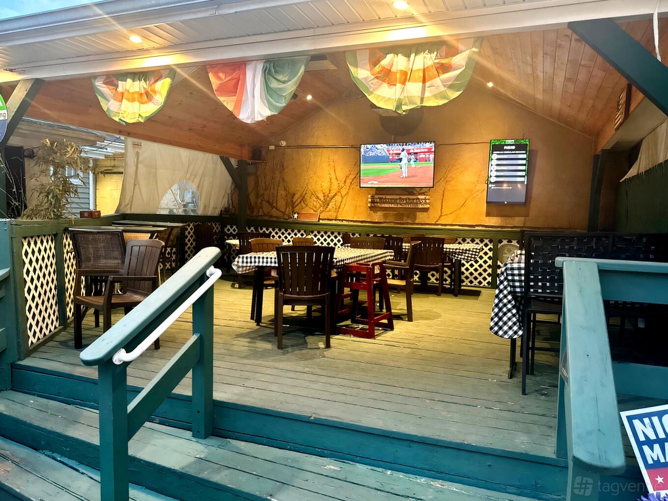 A pub deck room with wooden tables, checkered tablecloths, mounted TV, and Irish flag bunting at Joyce's Tavern.