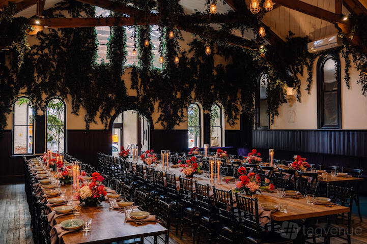 A function room with long wooden tables, black chairs, floral centerpieces, and greenery hanging from ceiling beams at The Hall at Welcome to Brunswick.