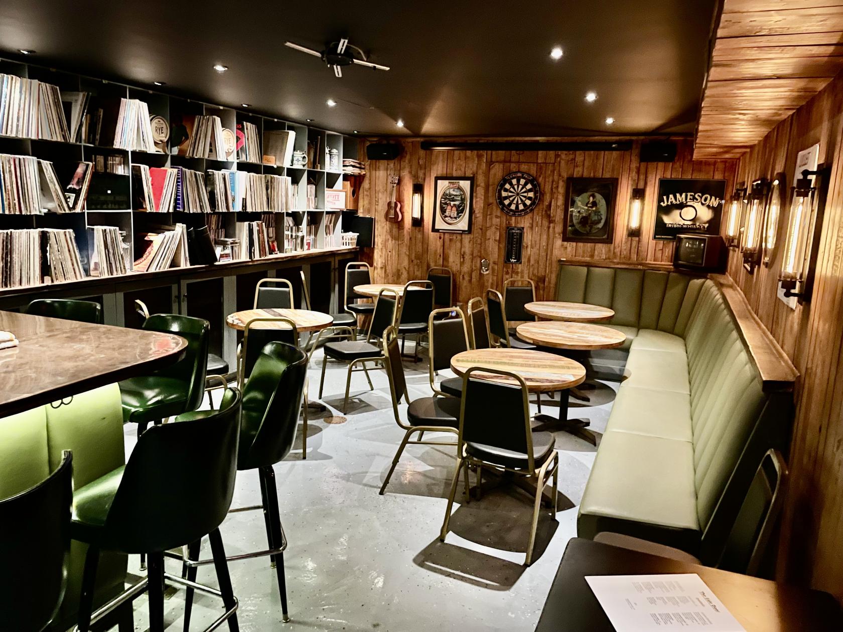 A function room in a pub with wood-paneled walls, round tables, green chairs, and shelves filled with vinyl records at The Pub and The People.