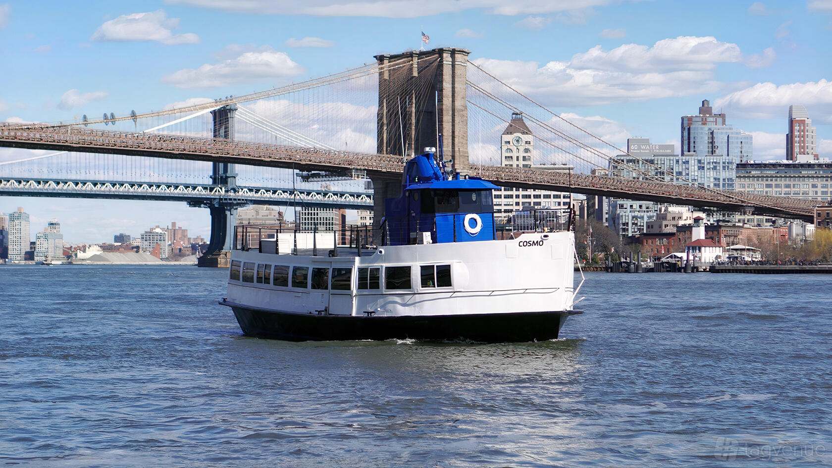 A boat with a white exterior and blue trim cruising on the river near the Brooklyn Bridge at Empire Cruises Cosmo.