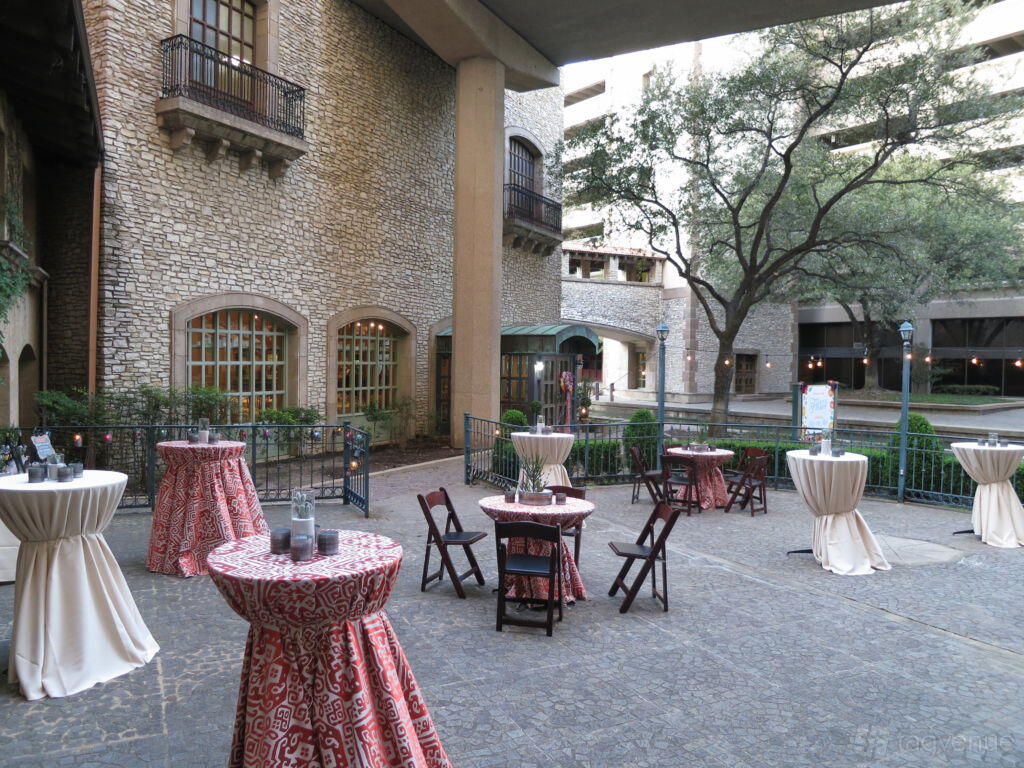 A hall courtyard with cocktail tables covered in patterned linens, folding chairs, stone walls, and mature trees at The Venetian Terrace.
