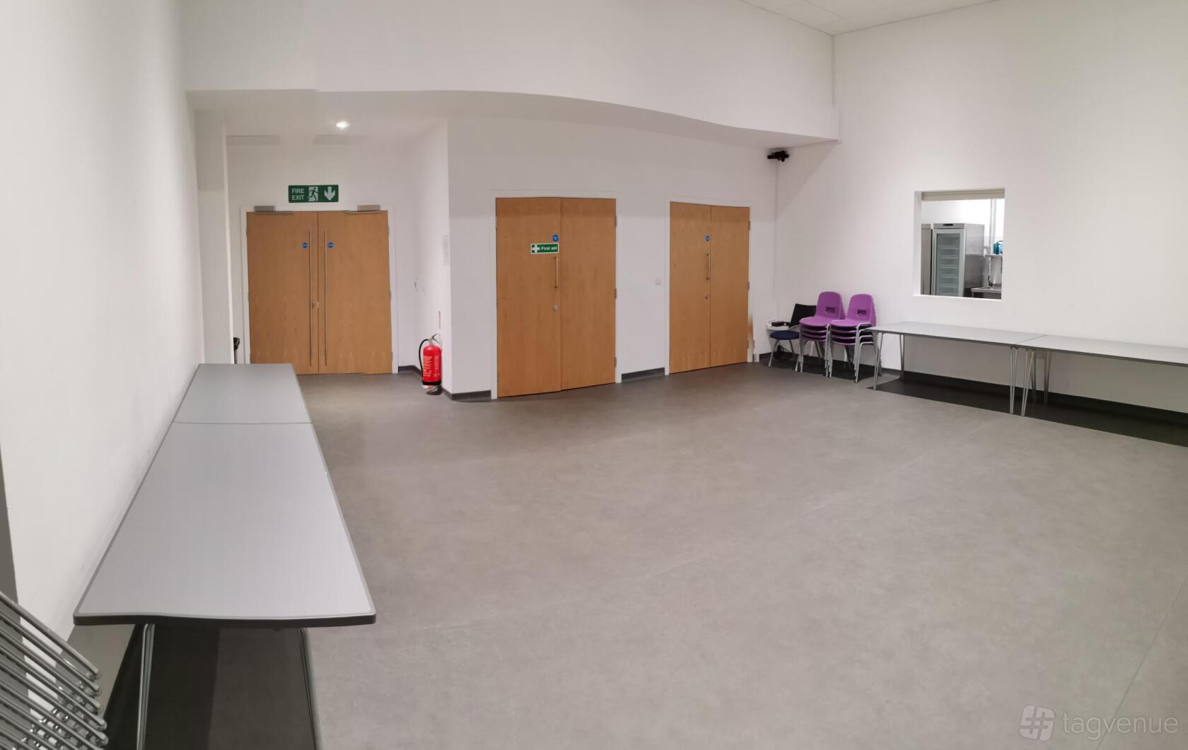 A hall with light grey flooring, white walls, folding tables, stacked chairs, and wooden doors at Victoria Hall.