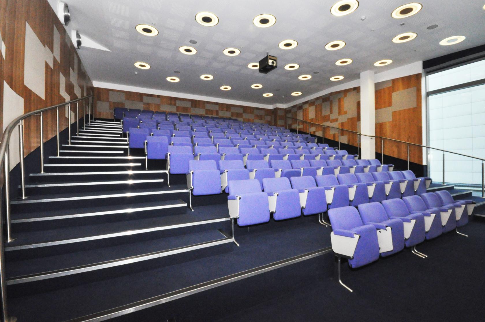 A university lecture theatre with tiered rows of blue seats, wood panel walls, and overhead circular lighting at Paragon House.