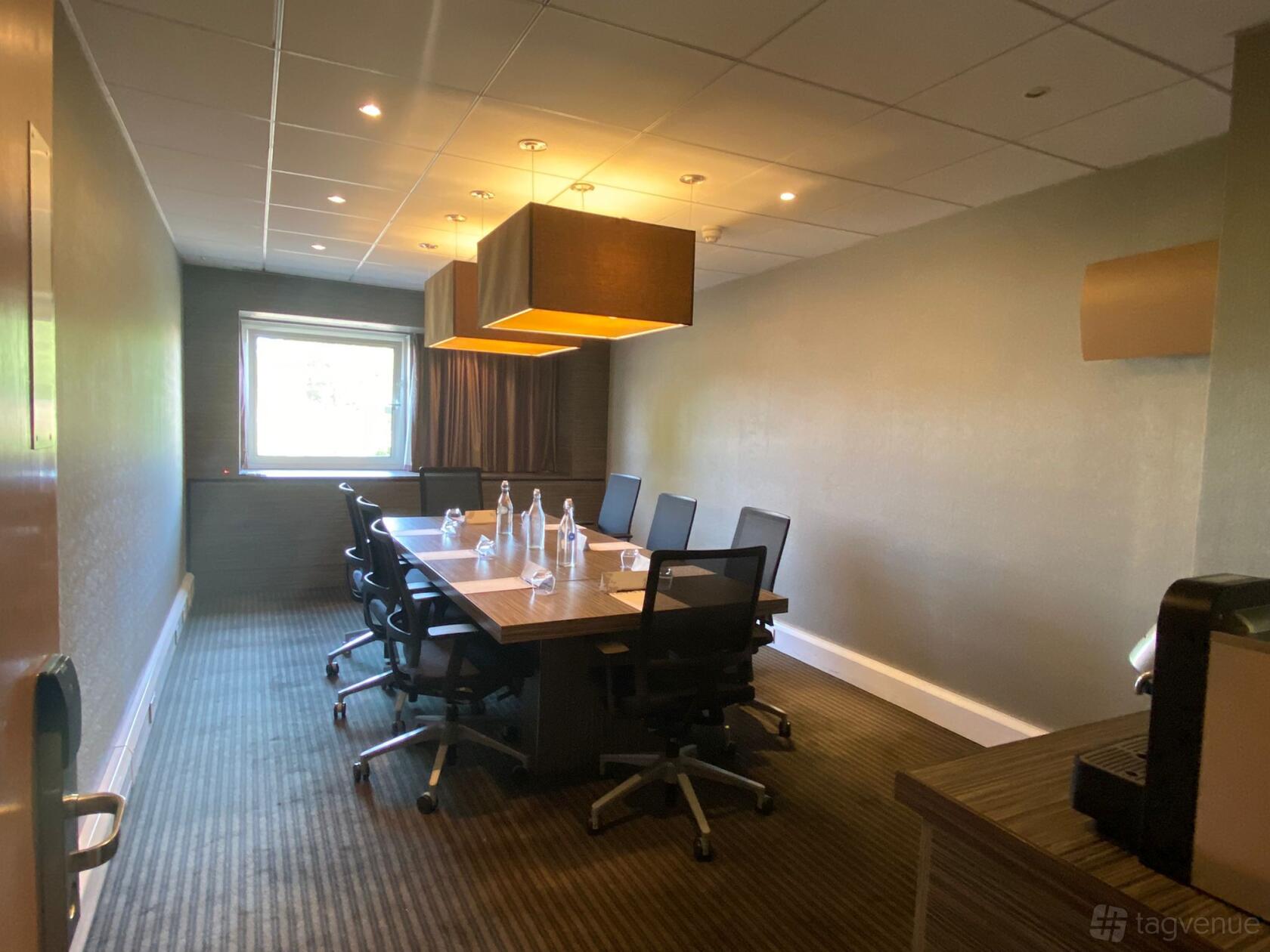A meeting room with a long wood table, black office chairs, ceiling light fixtures, and a window at Novotel London Waterloo.