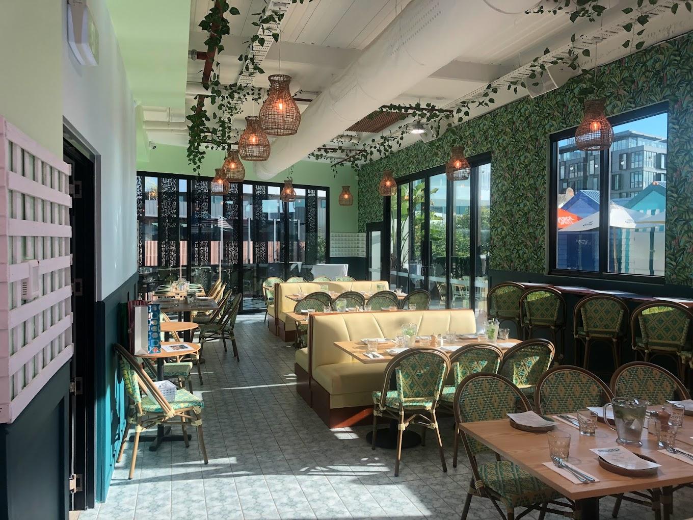 A bar with large windows, rattan pendant lights, patterned wallpaper, and banquette seating at The Bungalow Beer Garden.