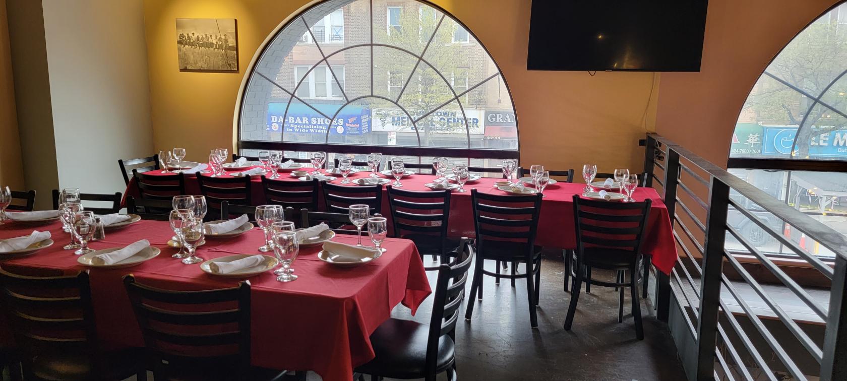 A restaurant mezzanine with long tables draped in red linens, set with glassware and napkins by an arched window at Patrizia's of Maspeth.
