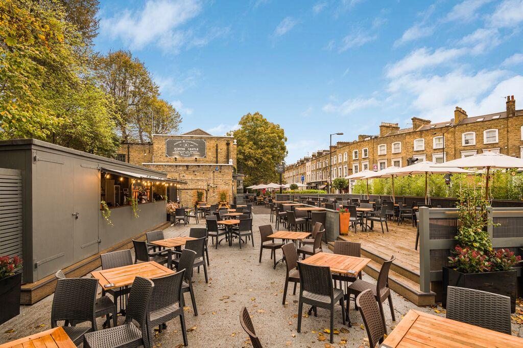 An outdoor pub garden with wooden tables, wicker chairs, and large umbrellas at The Alwyne Castle.