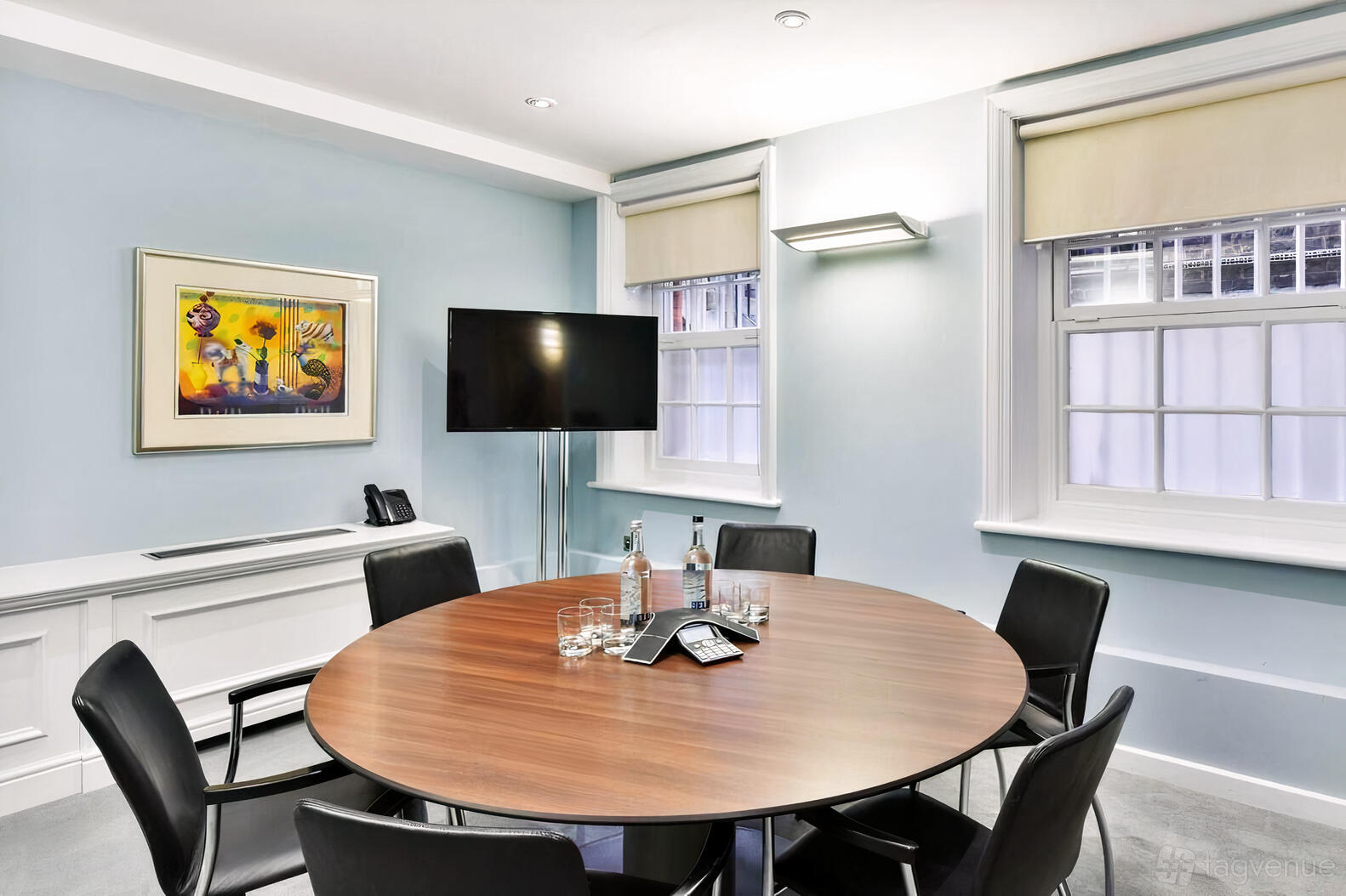 A meeting room with a round wooden table, black chairs, a wall-mounted TV, and large windows at 45 Pont Street.