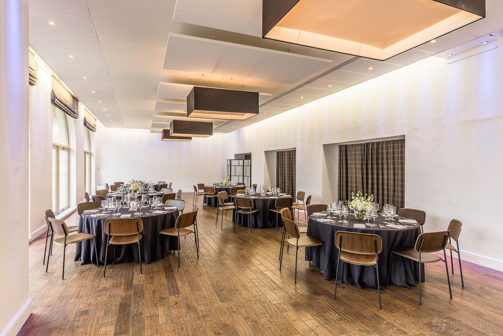 An event space in a hall with round tables set with glassware, wood flooring, and large windows at Glaziers Hall Limited.