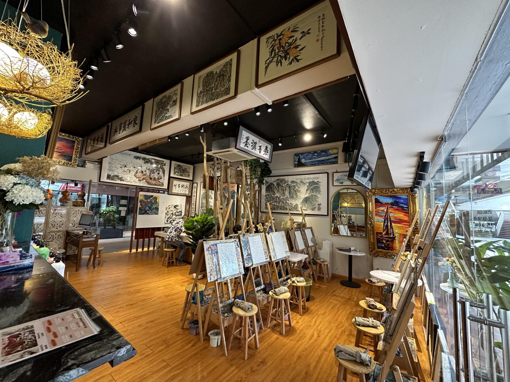 A gallery with easels, wooden flooring, framed artworks on the walls, and large windows at Sims Galleria.