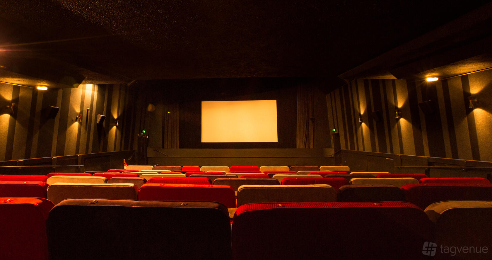 An auditorium with tiered cushioned seating, wall sconces, and a large projection screen at Everyman Gerrards Cross.