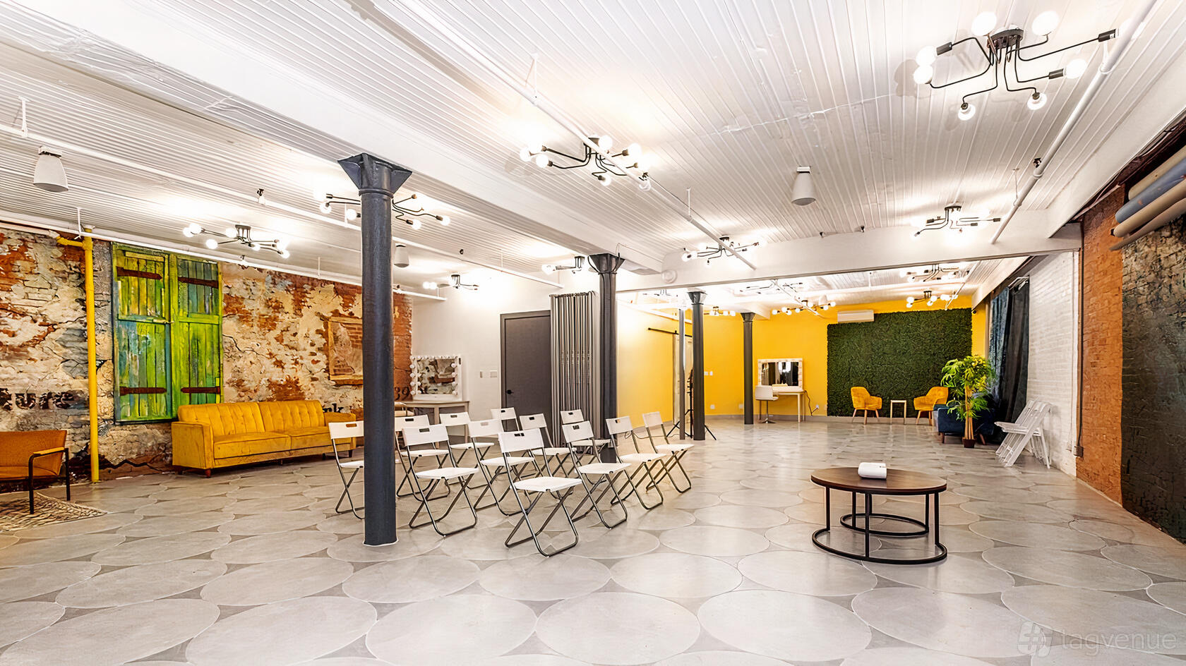 A photo studio with folding chairs, exposed brick, tall windows, and yellow accent walls at Studio Exhibit.