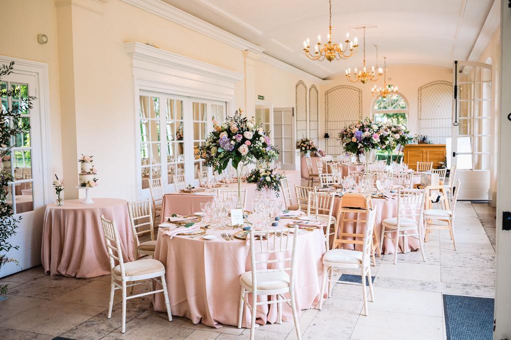 An event space with round tables draped in blush linens, floral arrangements, and chandeliers at Goldney House Orangery.