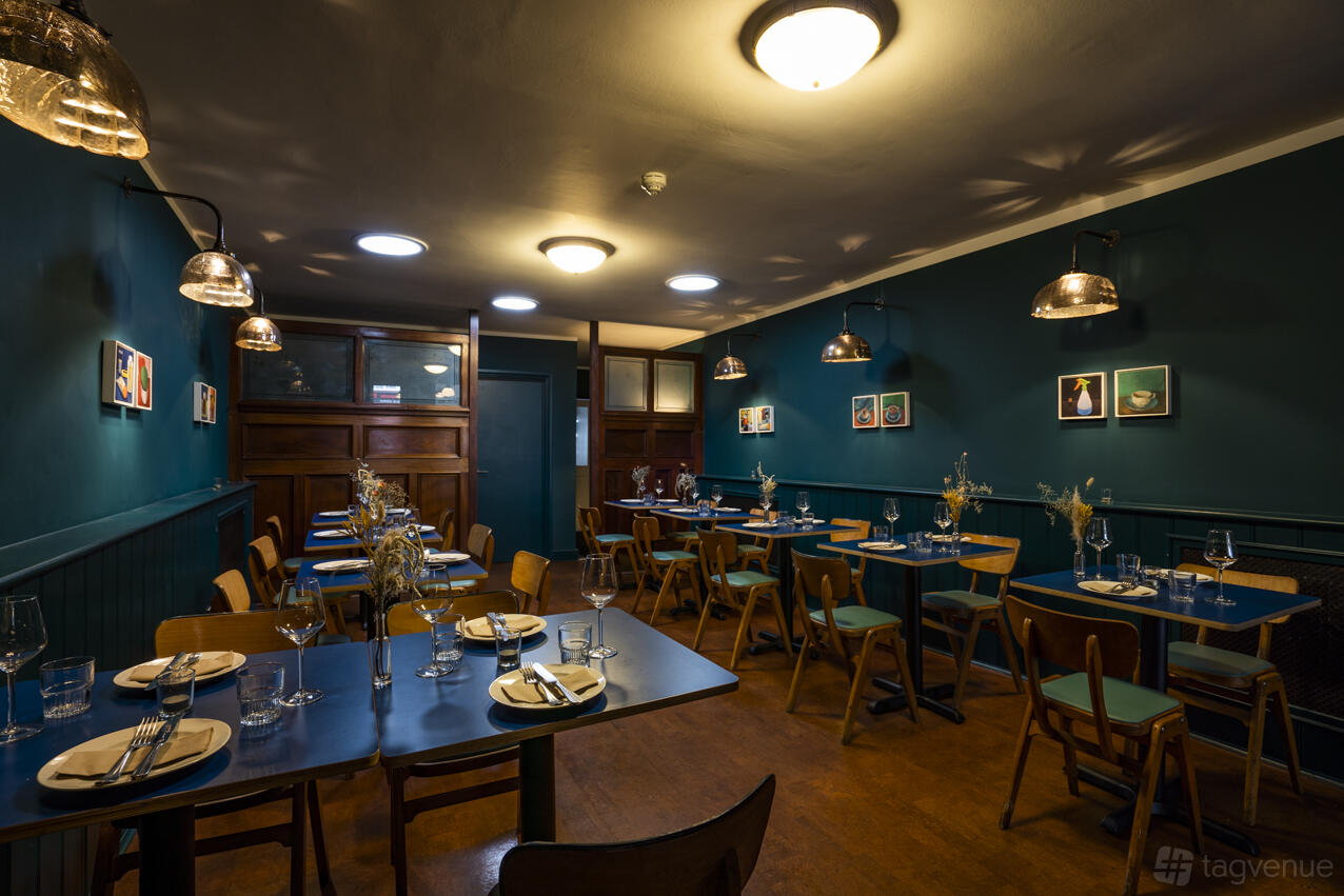 A restaurant with teal-painted walls, framed art, and wooden tables set with glassware at Klose & Soan.